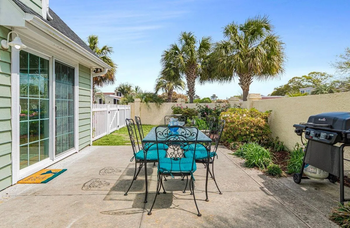This image spotlights a casual outdoor dining setup with a dining table and teal chairs, a grill ready for use, and palm trees within a fenced yard. The ambiance is relaxed and welcoming, ideal for alfresco meals or lounging while the kids and pets play close by.