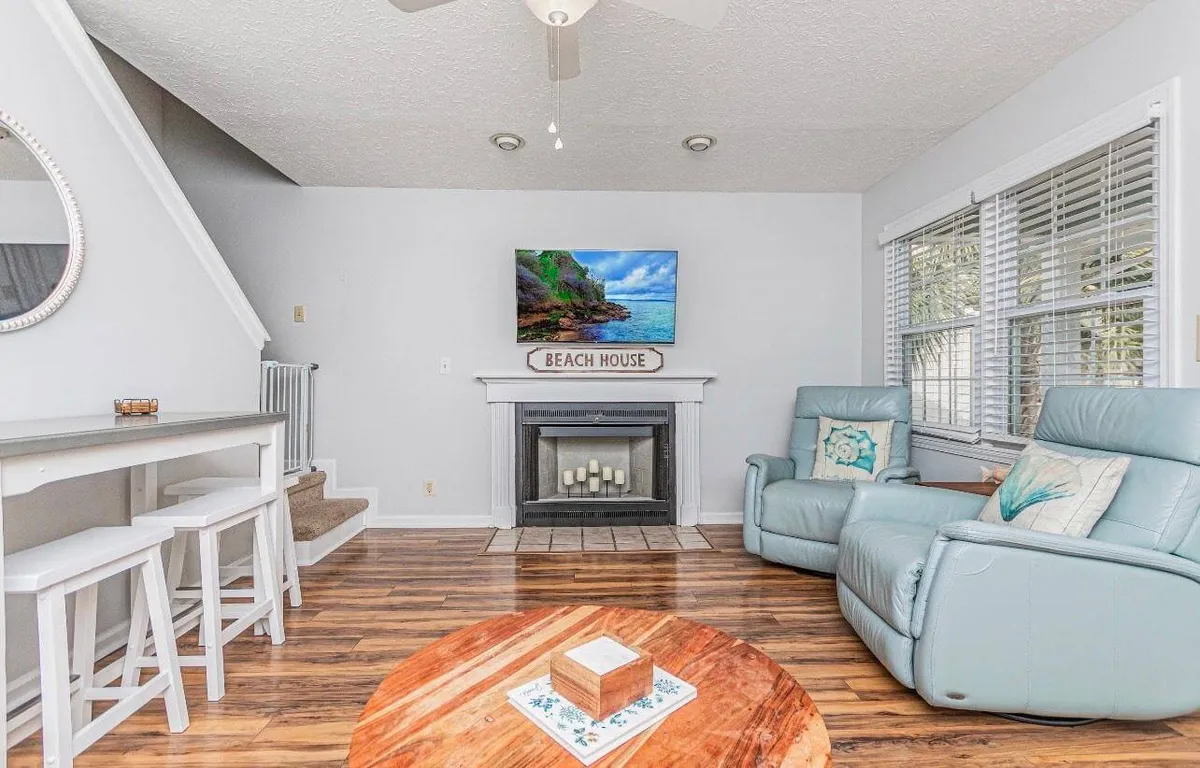 This image captures a comfortable living room area featuring plush seating, a round wooden coffee table, beach-themed decor, and a television displaying a scenic beach image. Sunlight streams into the space, creating a bright and inviting atmosphere ideal for relaxation and social gatherings.