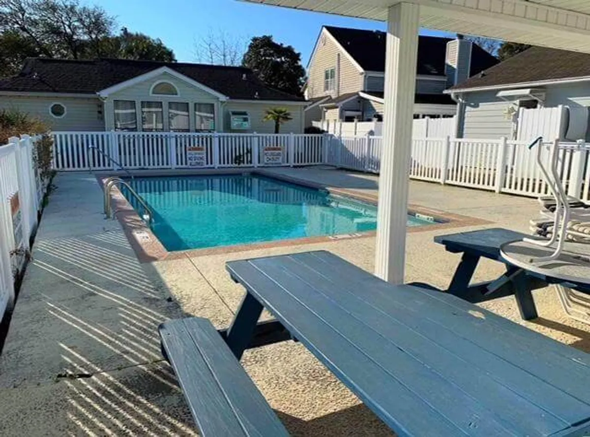 A sparkling swimming pool is the centerpiece, set within a fenced area with ample outdoor seating and a nearby house visible in the background. The mood is relaxed, suggesting leisurely summer days spent swimming, sunbathing, or hosting poolside gatherings.