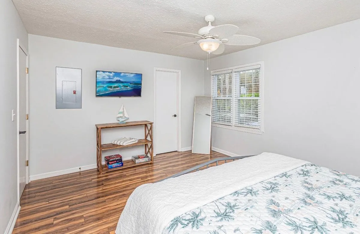 A cozy guest bedroom with a floral quilted bed, wooden shelving adorned with decorative accents, a framed seascape painting, a full-length mirror, and windows with blinds. The natural light and thoughtful decor create a warm and welcoming sanctuary.