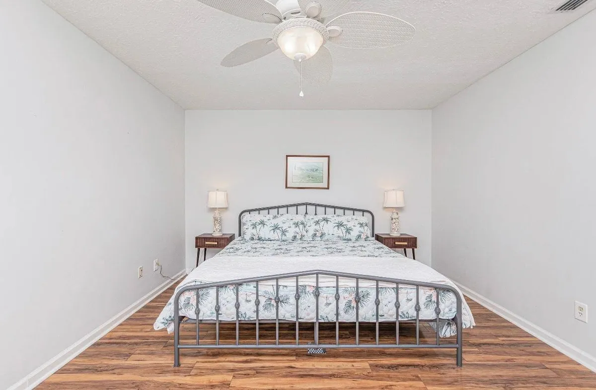 A serene bedroom featuring a plush bed with crisp white bedding, nightstands with matching lamps on either side, a ceiling fan overhead, and soothing wall art. The room is softly lit, evoking a calm and restful atmosphere ideal for unwinding after a busy day.