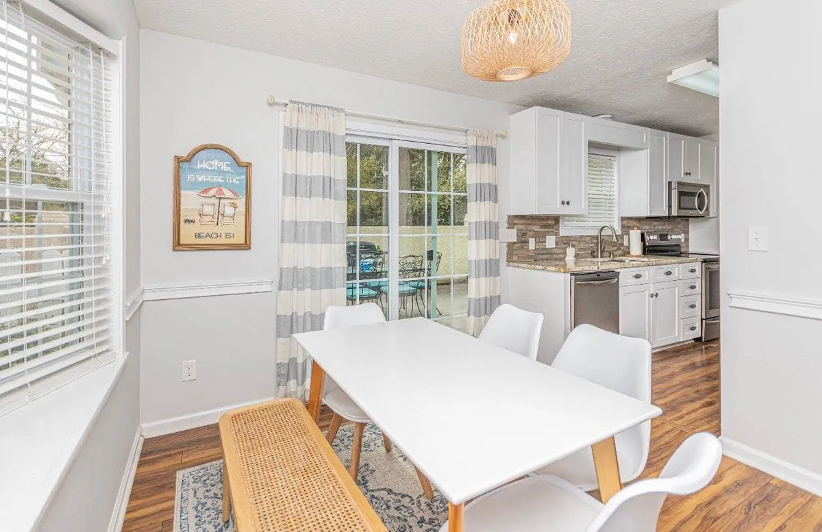 A bright and welcoming dining room and kitchen area with a large dining table, kitchen in the background, expansive windows, and a stylish light fixture overhead. The seamless layout encourages easy flow between cooking, dining, and socializing.