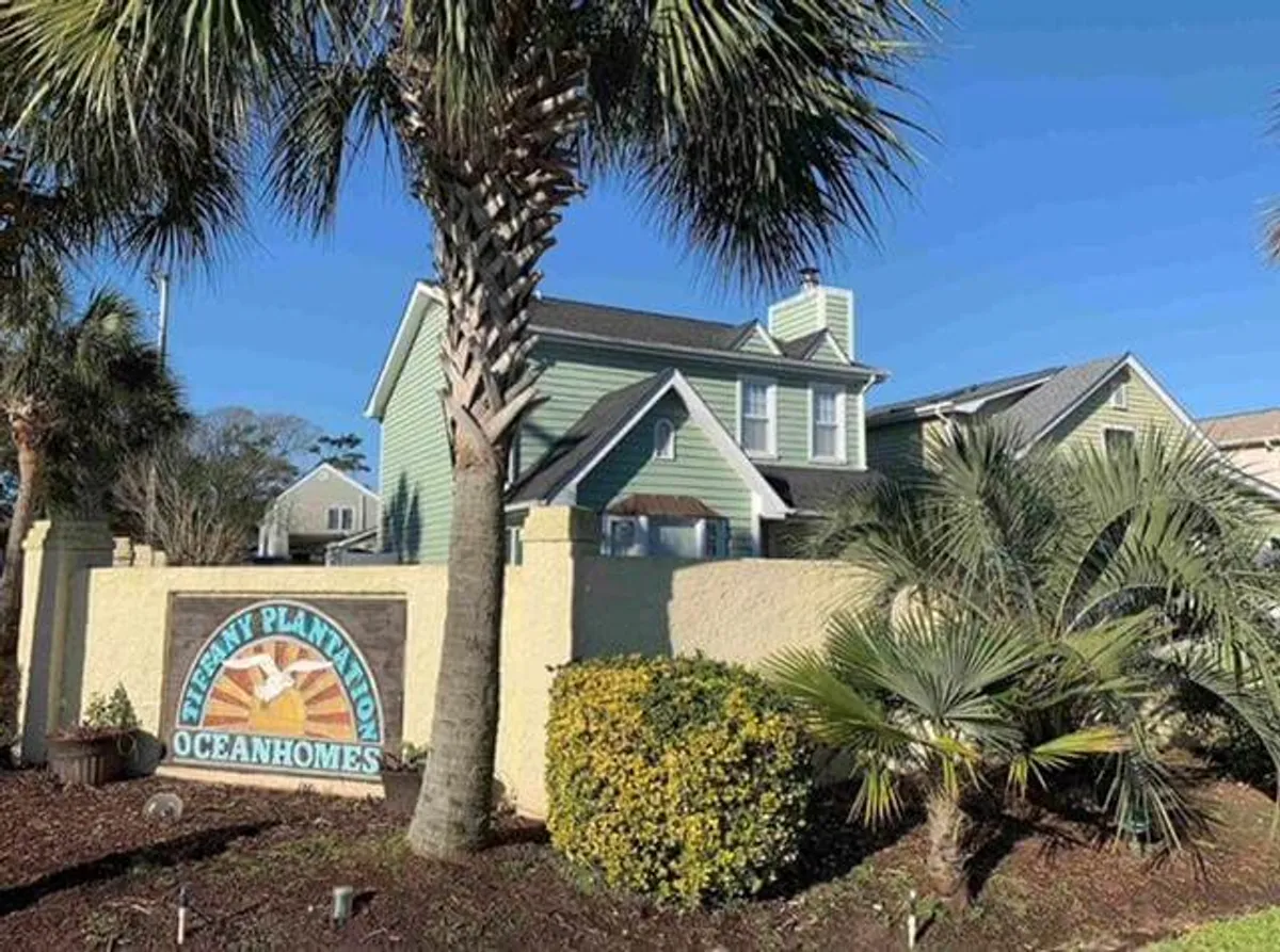 A broad exterior shot of the property’s green house, framed by palm trees and attractive landscaping. Visible signage and open surroundings evoke a sense of arrival and connection to the neighborhood, suggesting proximity to the beach and local amenities. The inviting mood hints at outdoor relaxation and community activities just steps from the home.
