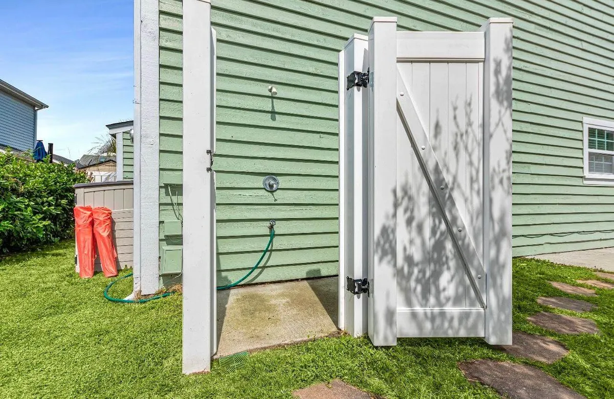 An exterior view featuring a welcoming outdoor shower set beside a green-sided wall and pathway stones. Red towels hang ready for use, and lush greenery creates a relaxed transition between indoor comfort and outdoor beach adventures. The space is sunlit and feels like an essential stop after sandy, fun-filled excursions.