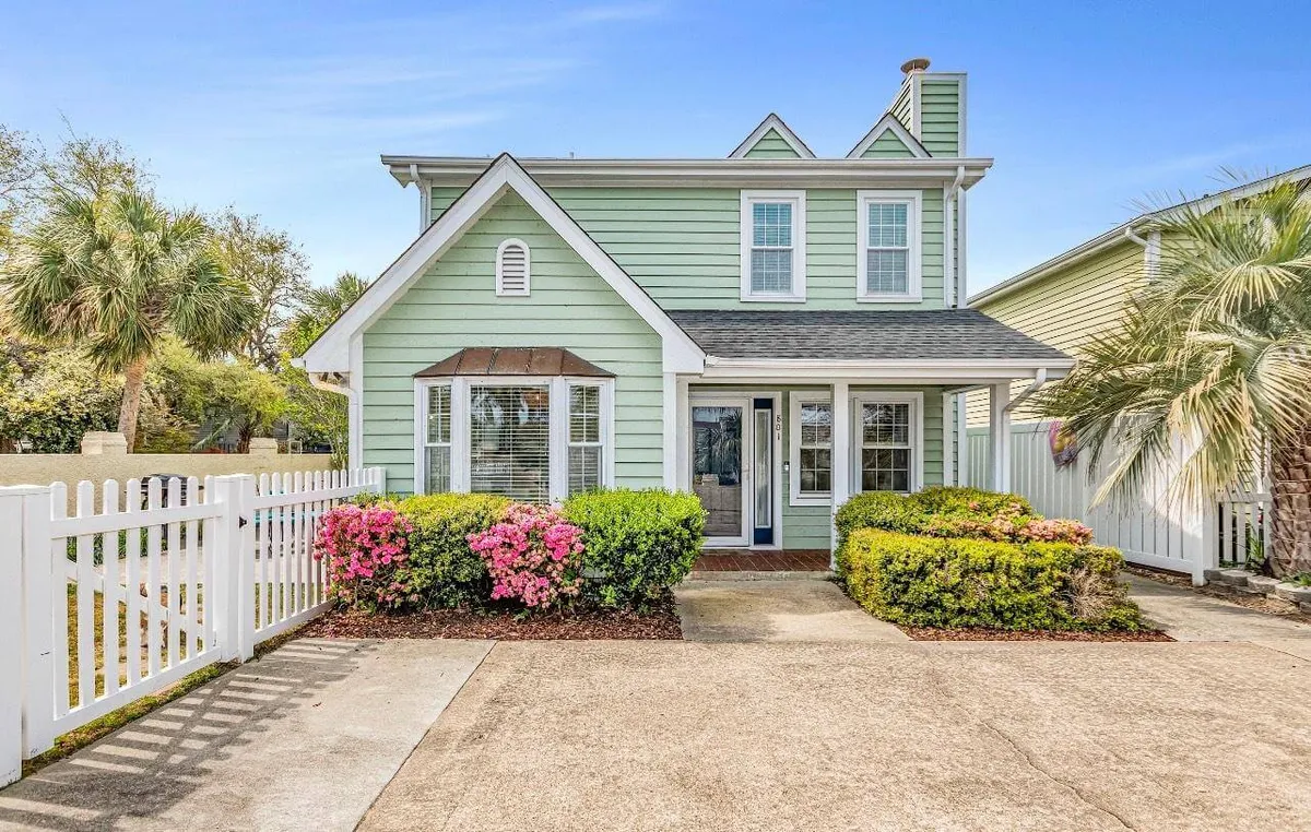 The image shows the cheerful green exterior of a charming single-family home in North Myrtle Beach, framed by blooming flowers and a classic white picket fence. The inviting front porch is perfect for relaxing, while the lush yard hints at a welcoming, pet-friendly atmosphere.