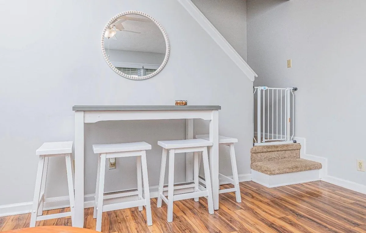 The image features a bright, inviting breakfast bar with a white bar table, matching stools, and a round mirror reflecting soft natural light. The setup encourages casual dining, morning coffee, or quick snacks with friends.